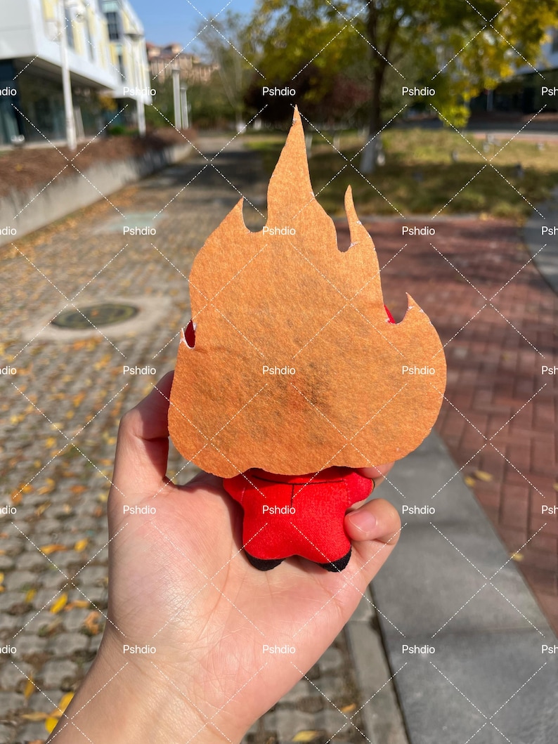 May include: A plush toy shaped like a flame, with an orange felt flame and a red body. The toy is held in a hand, with the background showing a blurred outdoor scene.