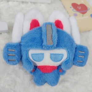 May include: A blue robot plush toy with white bunny ears and red details. The robot has a silver stripe on its head, blue eyes, and a red scarf. The toy is made of soft, fluffy material.