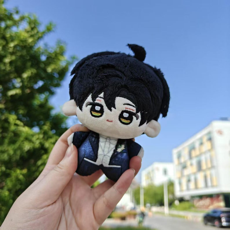 May include: A small plush doll with black hair and a dark blue suit. The doll has large, expressive eyes and a small, smiling mouth. The doll is held in a hand against a background of green trees and a blue sky.