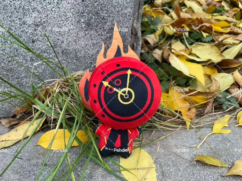 May include: A plush toy in the shape of a clock-faced character with a red and black design, yellow clock hands, and orange flames. The character is wearing a red suit with a black tie. The toy is surrounded by grass and yellow leaves.