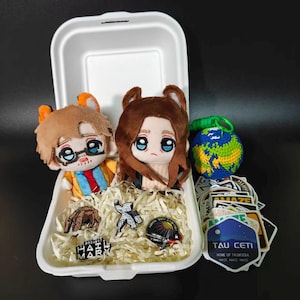 May include: A white takeaway container filled with shredded paper, two plush dolls with large blue eyes, a crocheted globe, and several stickers. The stickers feature text including Project Hail Mary and Tau Ceti, Home of Taumoeba, Amaze Amaze Amaze.