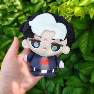 May include: A plush doll featuring a character with black hair, a white streak, and blue eyes. The doll wears a dark blue outfit with a red emblem and a black belt. The doll is held against a backdrop of green foliage.