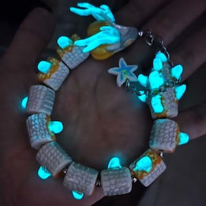 Pode incluir: Um bracelete que brilha no escuro com contas brancas e amarelas, com um pequeno amuleto de veado que brilha no escuro e um amuleto de estrela.