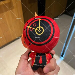 May include: A plush toy with a red circular head featuring a black and yellow clock design. The toy has a small orange fin and wears a red jacket with black and yellow accents. The toy is held in a person's hand.