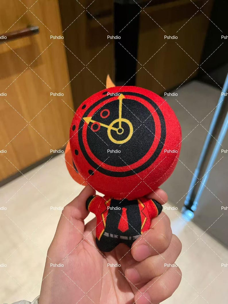 May include: A plush toy with a red circular head featuring a black and yellow clock design. The toy has a small orange fin and wears a red jacket with black and yellow accents. The toy is held in a person's hand.