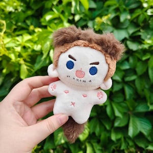 Pode incluir: Uma pequena boneca de pelúcia com corpo branco, cabelo castanho e olhos azuis. A boneca tem nariz e boca rosa, e pegadas rosas nas mãos e pés. Tem uma pequena cauda castanha.
