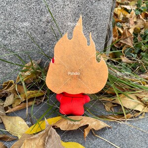 May include: A felt figure in the shape of a flame, with a red body and a tan flame-shaped head. The figure is set against a backdrop of fallen leaves and grass, creating a seasonal aesthetic.