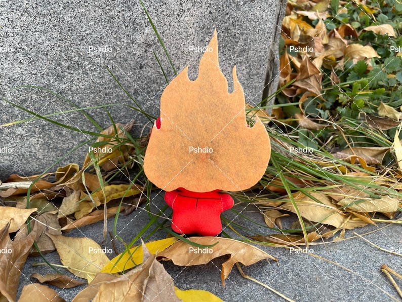 May include: A felt figure in the shape of a flame, with a red body and a tan flame-shaped head. The figure is set against a backdrop of fallen leaves and grass, creating a seasonal aesthetic.