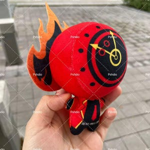 May include: A plush toy in the shape of a red character with a clock face on its head and flame-like accents. The toy has black details and is held in a hand.