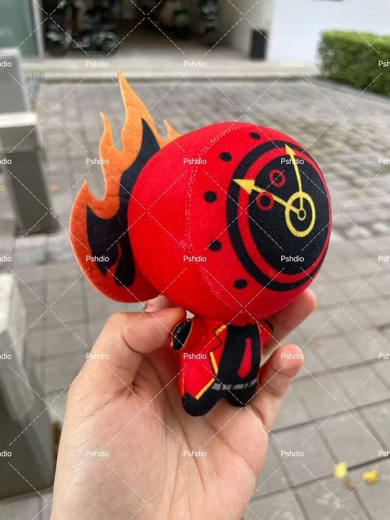 May include: A plush toy in the shape of a red character with a clock face on its head and flame-like accents. The toy has black details and is held in a hand.