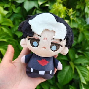 May include: A plush doll with black hair and a white streak, wearing a dark blue outfit with a red emblem. The doll has light blue eyes and a neutral expression. The doll is held against a backdrop of green foliage.