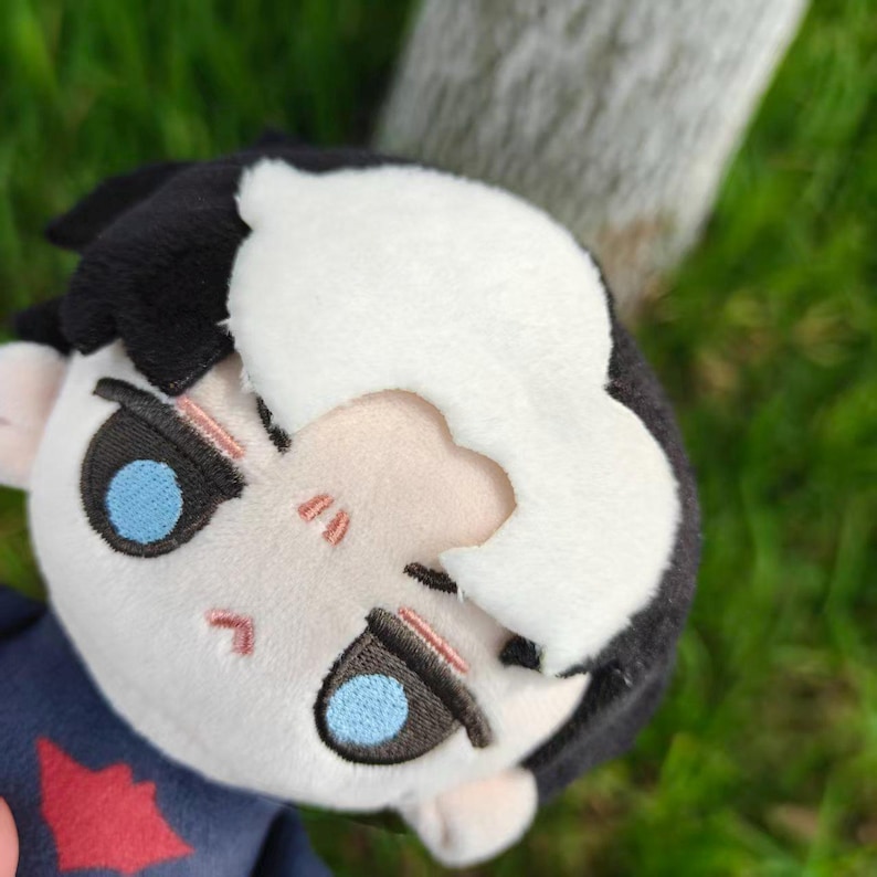 May include: A plush doll with a white and black hairstyle and blue eyes. The doll has a light skin tone and a red emblem on its dark blue shirt. The background is blurred green grass and a tree trunk.