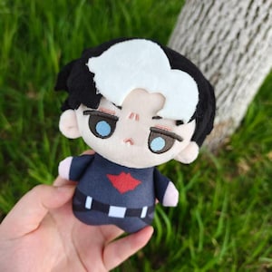 May include: A plush doll with black hair, a white streak, and blue eyes. The doll wears a navy blue outfit with a red emblem and a white belt. The doll is held against a background of green grass and a tree trunk.