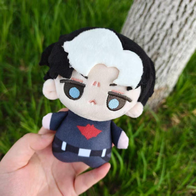 May include: A plush doll with black hair, a white streak, and blue eyes. The doll wears a navy blue outfit with a red emblem and a white belt. The doll is held against a background of green grass and a tree trunk.