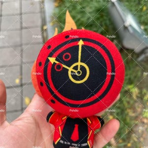 May include: A plush toy featuring a red and black circular design with yellow arrows and a central clock face. The toy has a red jacket with black lapels and a red tie. The toy is held in a hand.