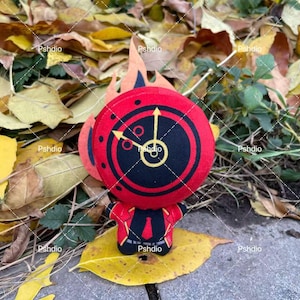May include: A plush toy featuring a red and black clock face design with yellow hands and accents. The toy has a small body and is set against a backdrop of autumn leaves and foliage.
