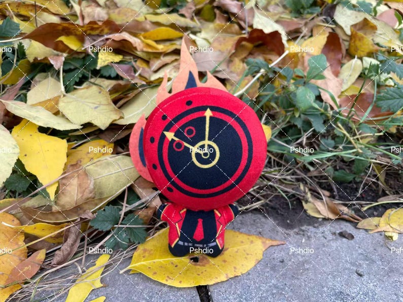 May include: A plush toy featuring a red and black clock face design with yellow hands and accents. The toy has a small body and is set against a backdrop of autumn leaves and foliage.