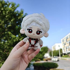 May include: A plush doll with a white suit, red vest, and black bow tie. The doll has white hair, large red eyes, and a small, embroidered face. The doll is held in a hand against a background of green trees and a blue sky.
