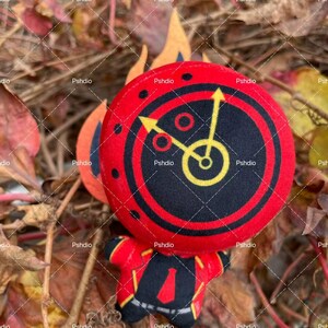 May include: A plush toy in the shape of a clock with a red and black face, yellow hands, and a red body. The clock face has a black center with yellow clock hands. The toy is set against a backdrop of fall foliage.