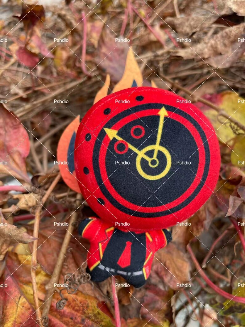 May include: A plush toy in the shape of a clock with a red and black face, yellow hands, and a red body. The clock face has a black center with yellow clock hands. The toy is set against a backdrop of fall foliage.