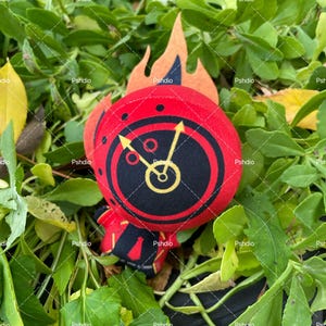 May include: A plush toy with a red circular design featuring a black center with a yellow clock face. The toy has a flame-shaped orange felt accent. The toy is set against a backdrop of green foliage.