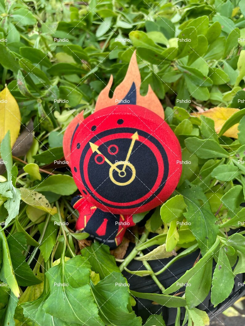 May include: A plush toy with a red circular design featuring a black center with a yellow clock face. The toy has a flame-shaped orange felt accent. The toy is set against a backdrop of green foliage.