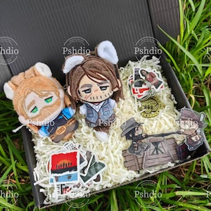 May include: A black gift box filled with plush dolls, stickers, and a keychain. The dolls are brown and tan with cartoon-like features. Stickers and a keychain feature characters and scenes. The box is lined with white paper shreds.