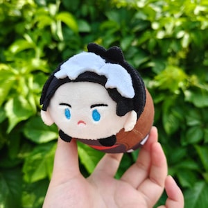 Peut inclure: Une poupée en peluche avec un visage de dessin animé, des cheveux noirs avec des reflets blancs et des yeux bleus. Le corps de la poupée est marron et beige. Elle est tenue dans une main devant un fond de feuillage vert.