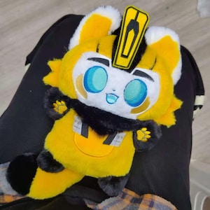 May include: A plush toy in the shape of a cartoon character, primarily yellow with black and white accents. It has large, embroidered blue eyes, a black collar, and a yellow emblem on its head. The toy is soft and cuddly.