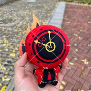 May include: A plush toy featuring a red circular head with a black clock design and yellow hands. The toy has a red body with black and yellow accents, and orange flame-shaped details. The toy is held in a hand.