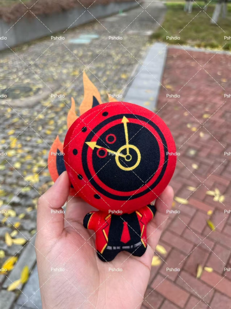 May include: A plush toy featuring a red circular head with a black clock design and yellow hands. The toy has a red body with black and yellow accents, and orange flame-shaped details. The toy is held in a hand.