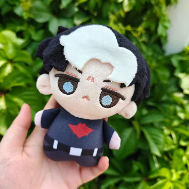 May include: A plush doll with black hair, a white streak, and blue eyes. The doll wears a navy blue outfit with a red emblem. The doll is held in a hand against a backdrop of green foliage.