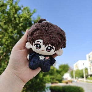 May include: A plush doll with brown hair and embroidered facial features. The doll is wearing a dark blue suit with a white shirt and a pink flower. The doll is held in a hand against a background of green foliage and a blue sky.