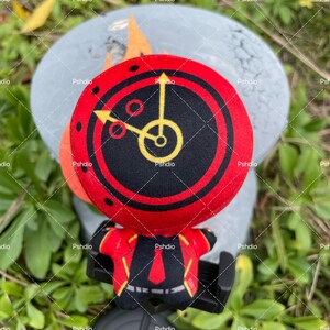 May include: A plush toy featuring a red and black clock face with yellow hands and accents. The toy has a matching red and black outfit. The background is a blurred gray surface and green foliage.