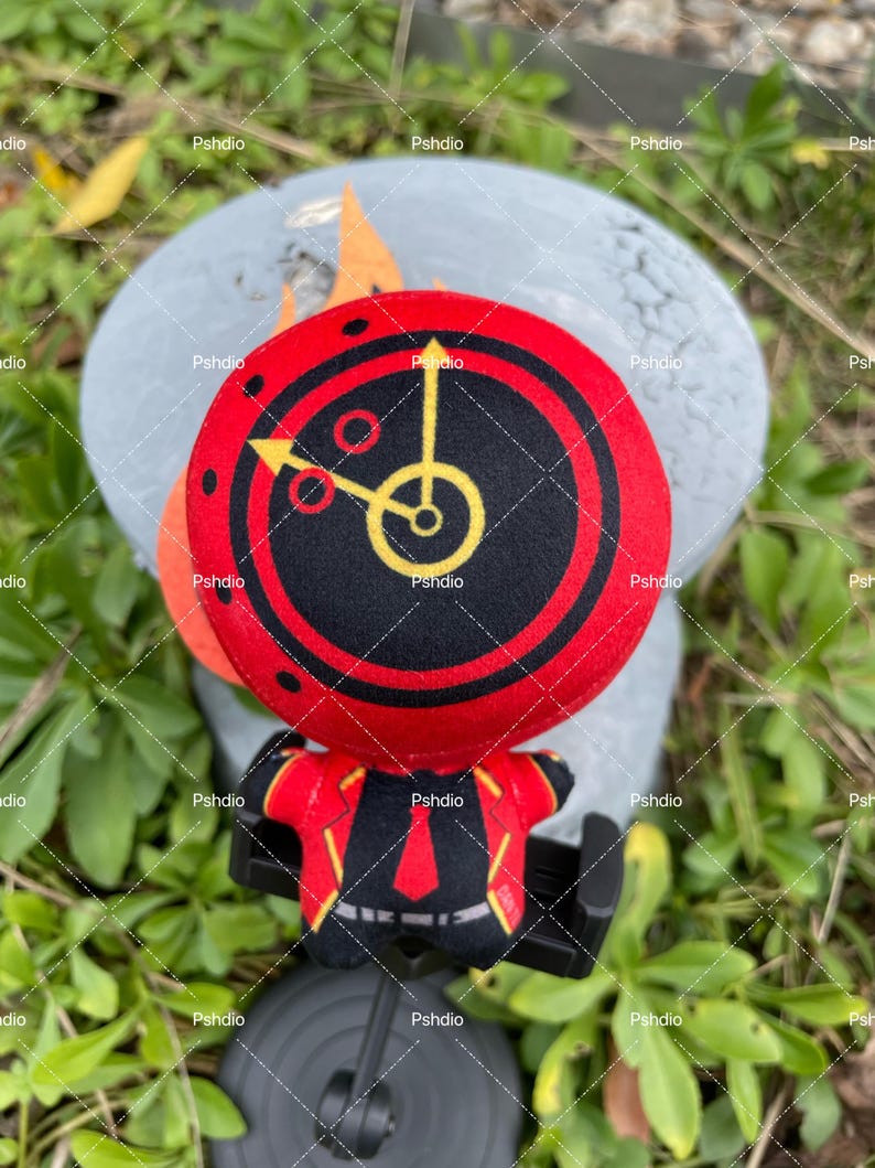 May include: A plush toy featuring a red and black clock face with yellow hands and accents. The toy has a matching red and black outfit. The background is a blurred gray surface and green foliage.