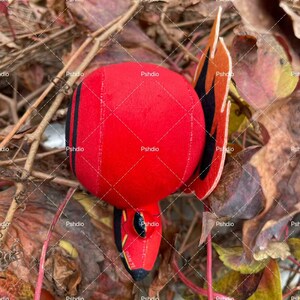 May include: A plush red bird toy with black stripes and accents. The bird has a round body and a pointed beak. The wings are orange and black. The toy is lying on a bed of brown and yellow leaves.