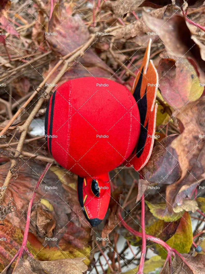 May include: A plush red bird toy with black stripes and accents. The bird has a round body and a pointed beak. The wings are orange and black. The toy is lying on a bed of brown and yellow leaves.