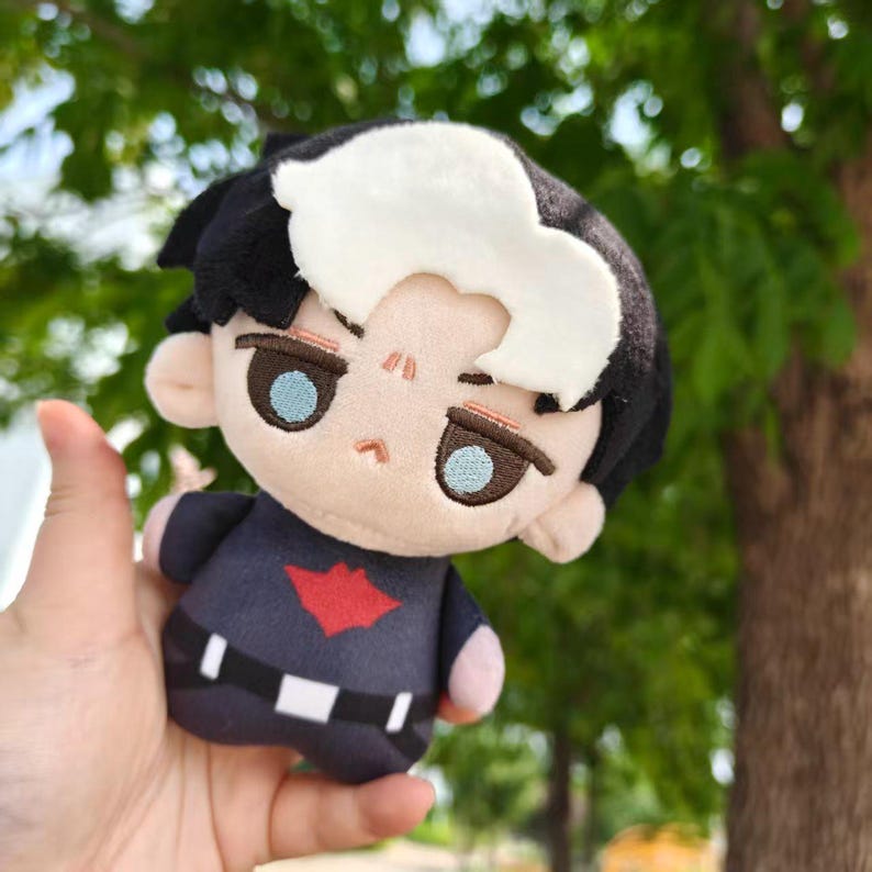 May include: A plush doll featuring a character with black hair, a white streak, and blue eyes. The doll wears a navy blue outfit with a red emblem. The doll is held in a hand against a blurred green background.