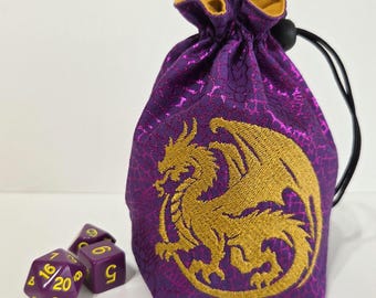 May include: A purple drawstring pouch with a gold embroidered dragon design on the front, featuring a black cord closure. Next to the pouch are two purple polyhedral dice with yellow numbers, including a twenty-sided die and a six-sided die.