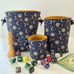 May include: Three navy blue drawstring dice bags with a celestial pattern of moons, suns, and stars, with yellow lining. Assorted dice and a chess piece are scattered in front of the bags. The bags are of different sizes.