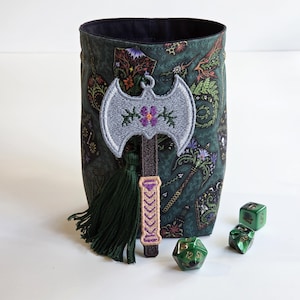 May include: A cylindrical fabric pouch featuring a dark green botanical and fantasy-themed print with a black interior. An embroidered silver double-bladed axe patch with a purple flower and a dark green tassel is attached to the front, alongside three green dice.