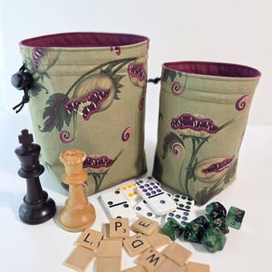 May include: Two drawstring dice bags with a botanical print of carnivorous plants in shades of green, purple, and cream. The bags are lined with a deep burgundy fabric. Chess pieces, dominoes, Scrabble tiles, and dice are scattered around the bags.