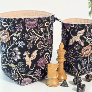 May include: Two fabric drawstring pouches featuring a black background with a floral and dragon pattern in shades of purple, peach, and cream. The pouches have a light beige interior lining and are displayed with wooden chess pieces and dice.