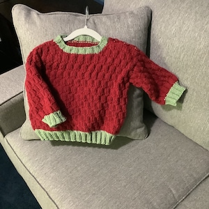 May include: A red knitted sweater with green trim. The sweater has a textured pattern and is hanging on a hanger.