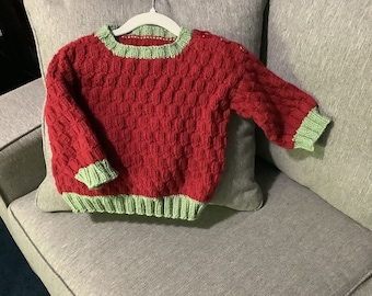 Könnte beinhalten: Ein roter Strickpullover mit grünem Besatz. Der Pullover hat ein strukturiertes Muster und hängt an einem Kleiderbügel.
