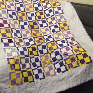 Könnte beinhalten: Eine Steppdecke mit einem karierten Muster in Violett-, Gelb- und Weißtönen. Die Steppdecke besteht aus Quadraten unterschiedlicher Größe und Farbe, die in einem zufälligen Muster angeordnet sind.