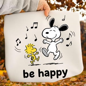 May include: A white tote bag featuring a black and white illustration of Snoopy and Woodstock dancing with musical notes. The text "be happy" is printed in black at the bottom of the bag. The bag is held by a person.