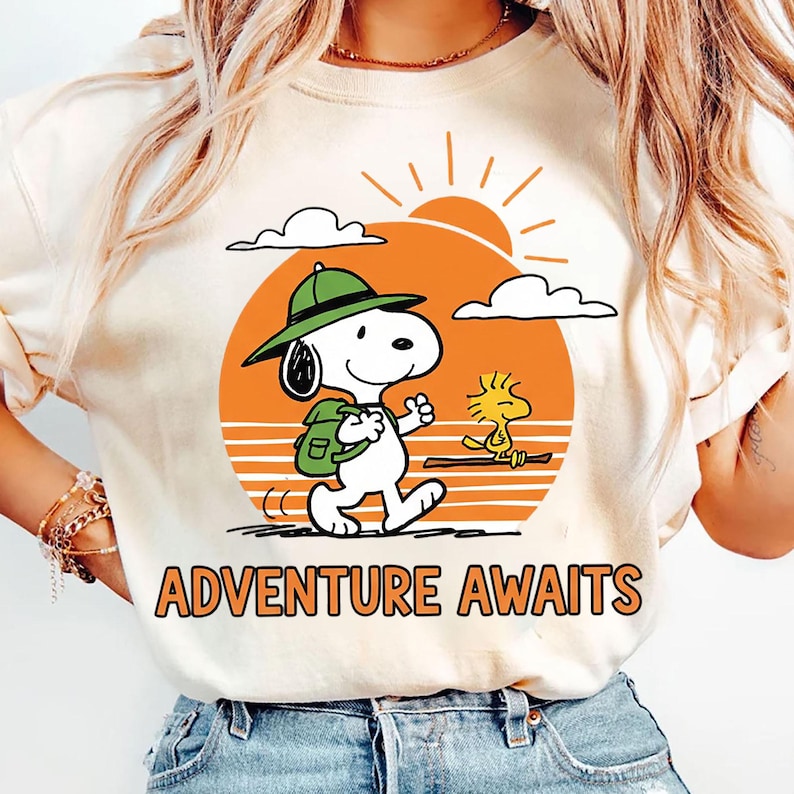 May include: Cream-colored t-shirt featuring a cartoon illustration of Snoopy with a backpack and a green hat, walking towards the right. The design includes an orange sun, clouds, and the text "ADVENTURE AWAITS" in orange.