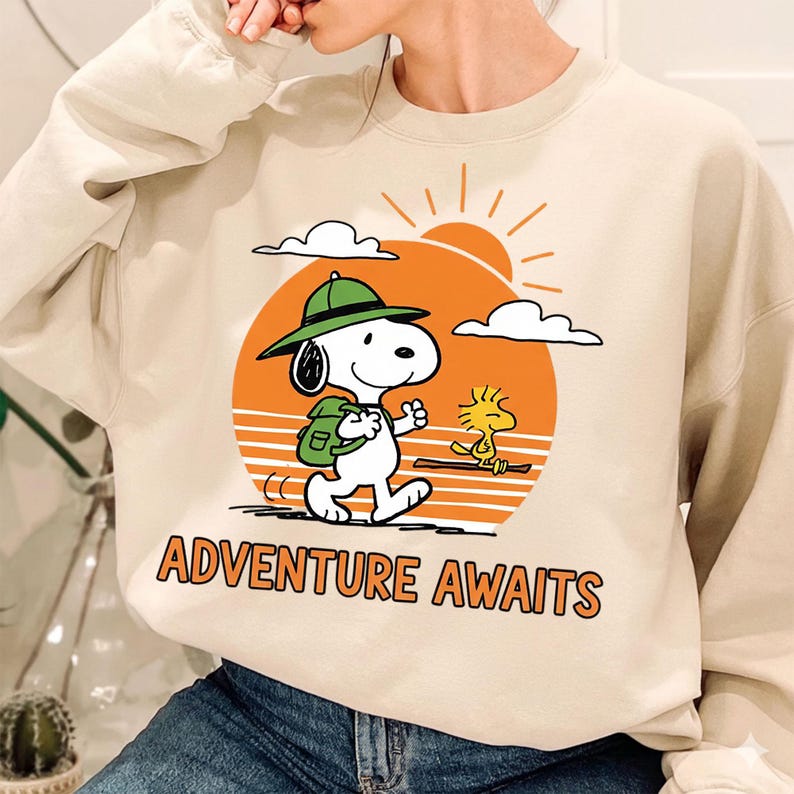 May include: Beige sweatshirt featuring a graphic of Snoopy with a green hat and backpack, walking towards the right. A yellow bird walks beside him. An orange sun and the words "ADVENTURE AWAITS" are also in the design.