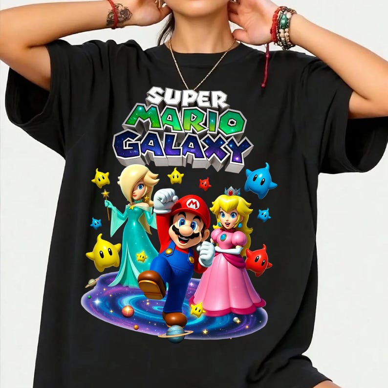 Puede incluir: Camiseta negra con un gr&aacute;fico colorido de Mario, la Princesa Peach y Rosalina de Super Mario Galaxy. El dise&ntilde;o incluye el texto "SUPER MARIO GALAXY" en 3D, con estrellas y un fondo de galaxia.